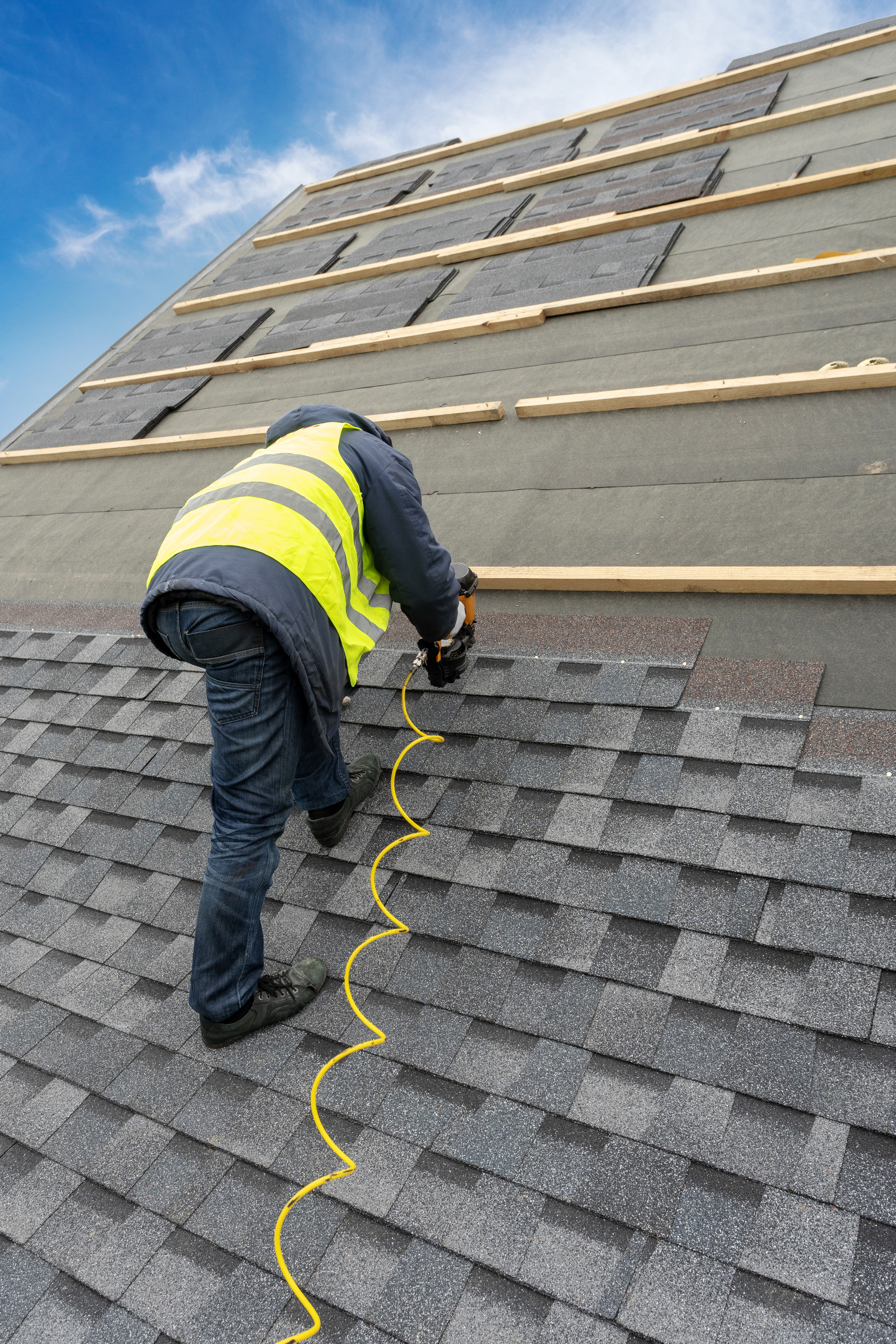 Vertical photo of unrecognizable roofer worker in uniform work wear using air or pneumatic nail gun and installing asphalt or bitumen tile on top of the roof under construction house